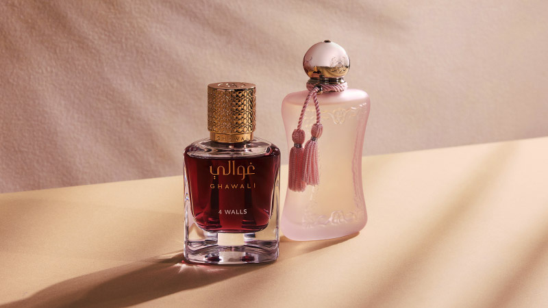 Two elegant perfume bottles on a soft beige surface. The left bottle is dark with a gold cap labeled "Ghawali." The right is pink, ornate, and tasseled.