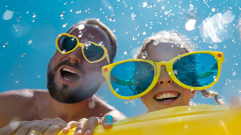 A man and a child wearing oversized yellow sunglasses are smiling and playing in water on a sunny day.